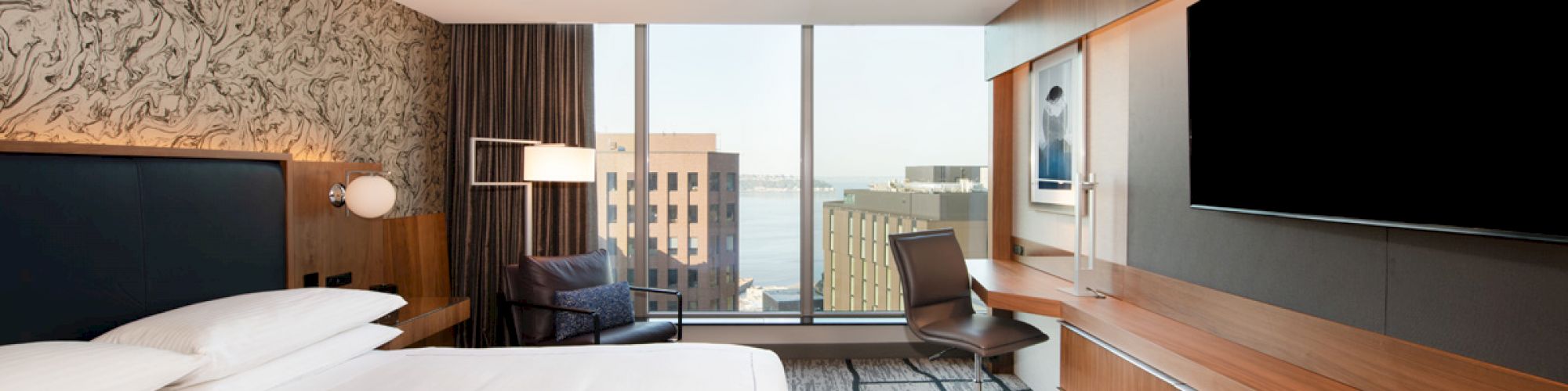 A modern hotel room with a large bed, a TV, desk, and chair, featuring a view of the city through a large window.