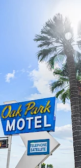 A motel sign reading "Oak Park Motel" with a blue sky, palm trees, and some buildings in the background. The sign also mentions telephone and satellite.