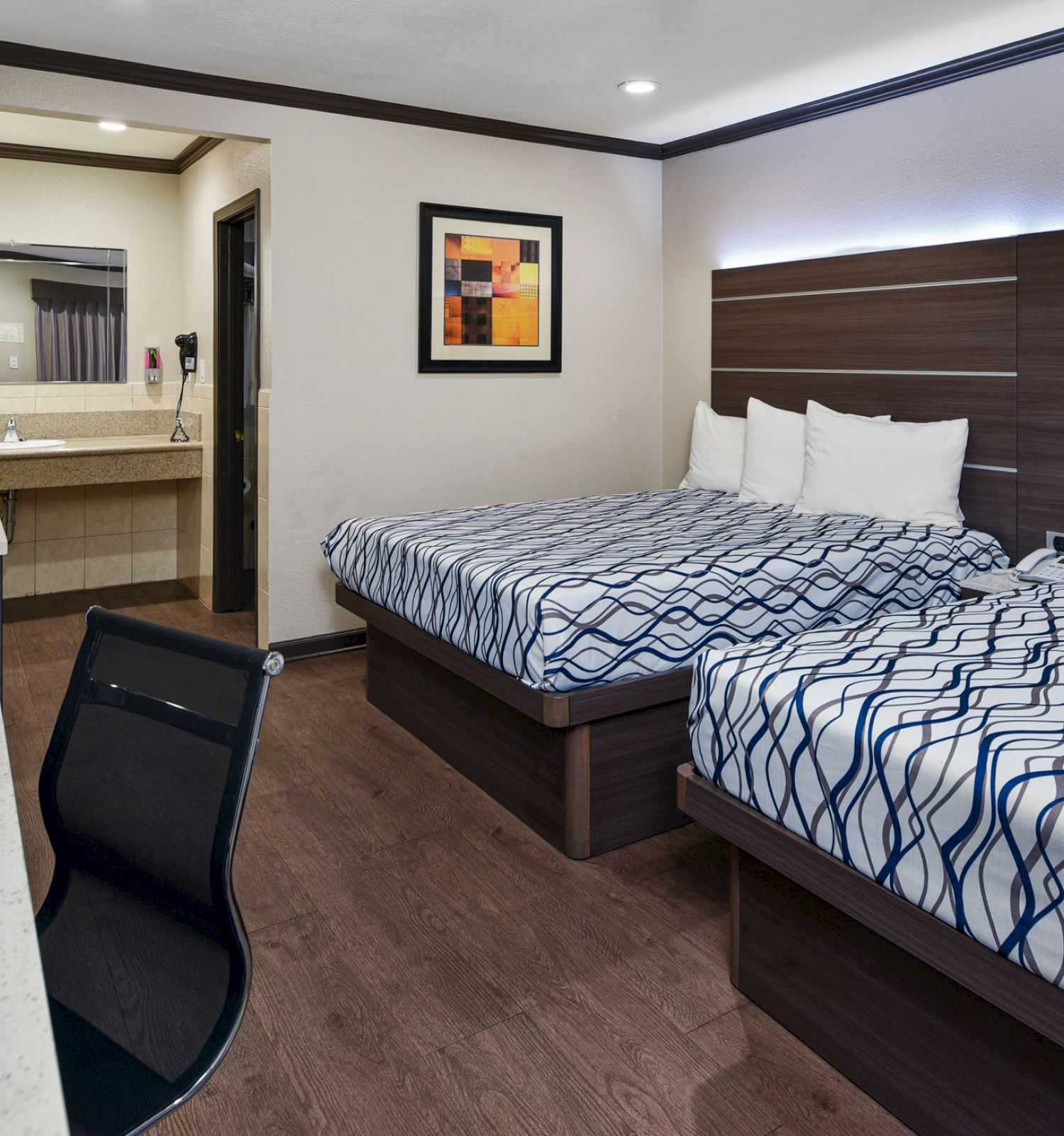 A modern hotel room features two double beds, a desk with a chair, and a dresser with a mirror. A bathroom is visible in the background.