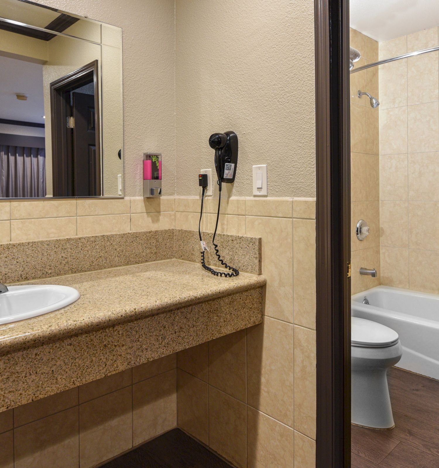 The image shows a bathroom with a large mirror, sink, countertop, wall-mounted hairdryer, and an adjoining bathtub with a shower and yellow curtain.