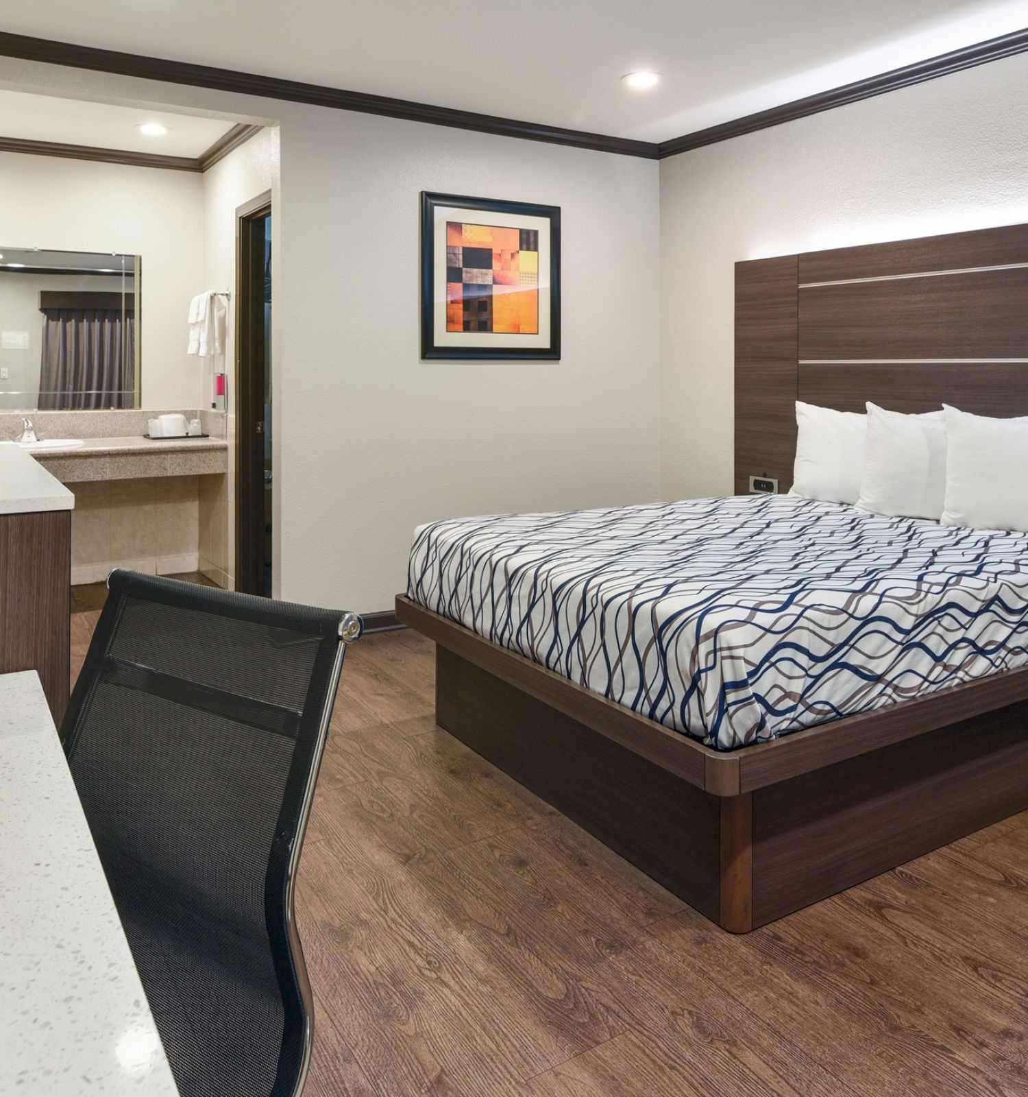 A modern hotel room with a bed, desk, chair, TV, and artwork. There's an attached bathroom visible through an open doorway.